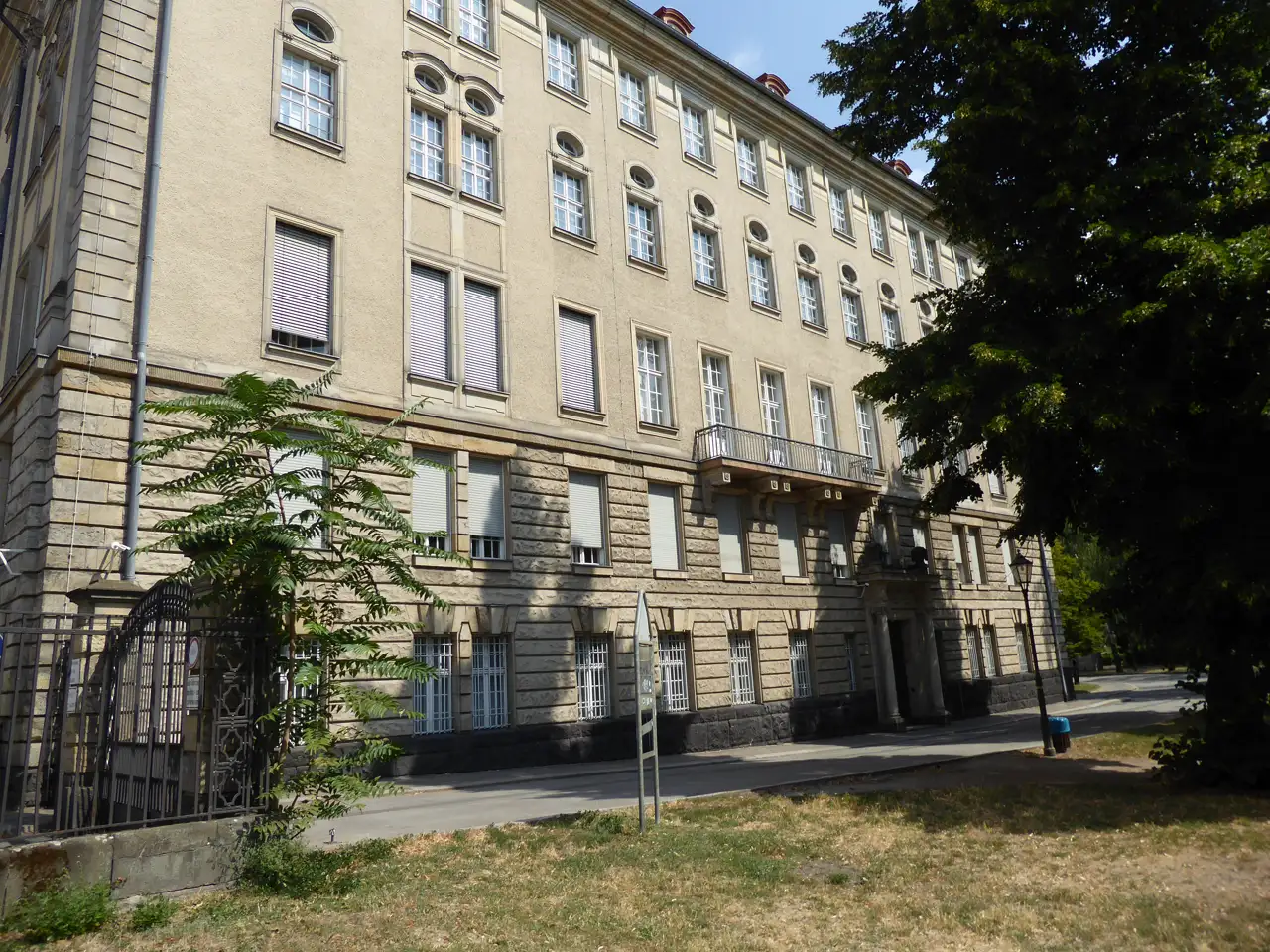 Fachada larga de piedra clara del edificio del Kammergericht, en la Elßholzstraße de Berlín-Schöneberg, con una verja, césped y árboles delante bajo un cielo despejado.