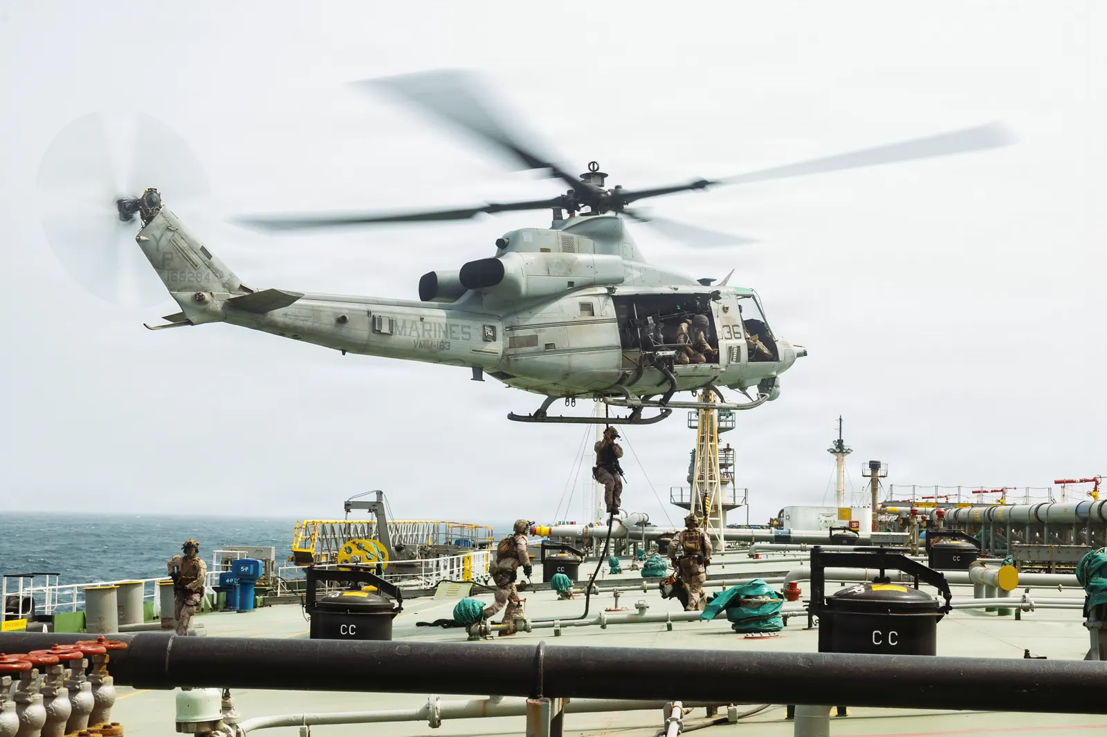 Un helicóptero UH-1Y de los Marines sobrevuela la cubierta de un petrolero mientras varios militares descienden por una cuerda y otros, ya en cubierta, avanzan entre las tuberías del barco.