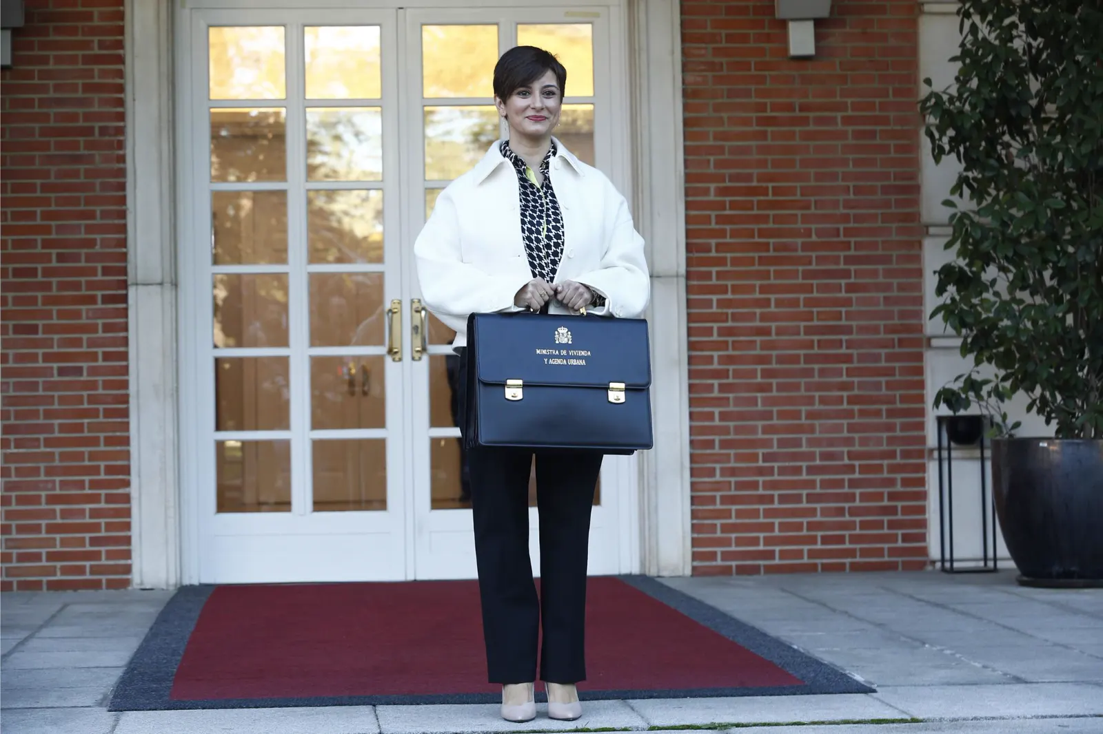 Isabel Rodríguez, de pie ante la puerta acristalada de un edificio de ladrillo, sostiene con las dos manos una cartera azul en la que se lee «Ministra de Vivienda y Agenda Urbana».