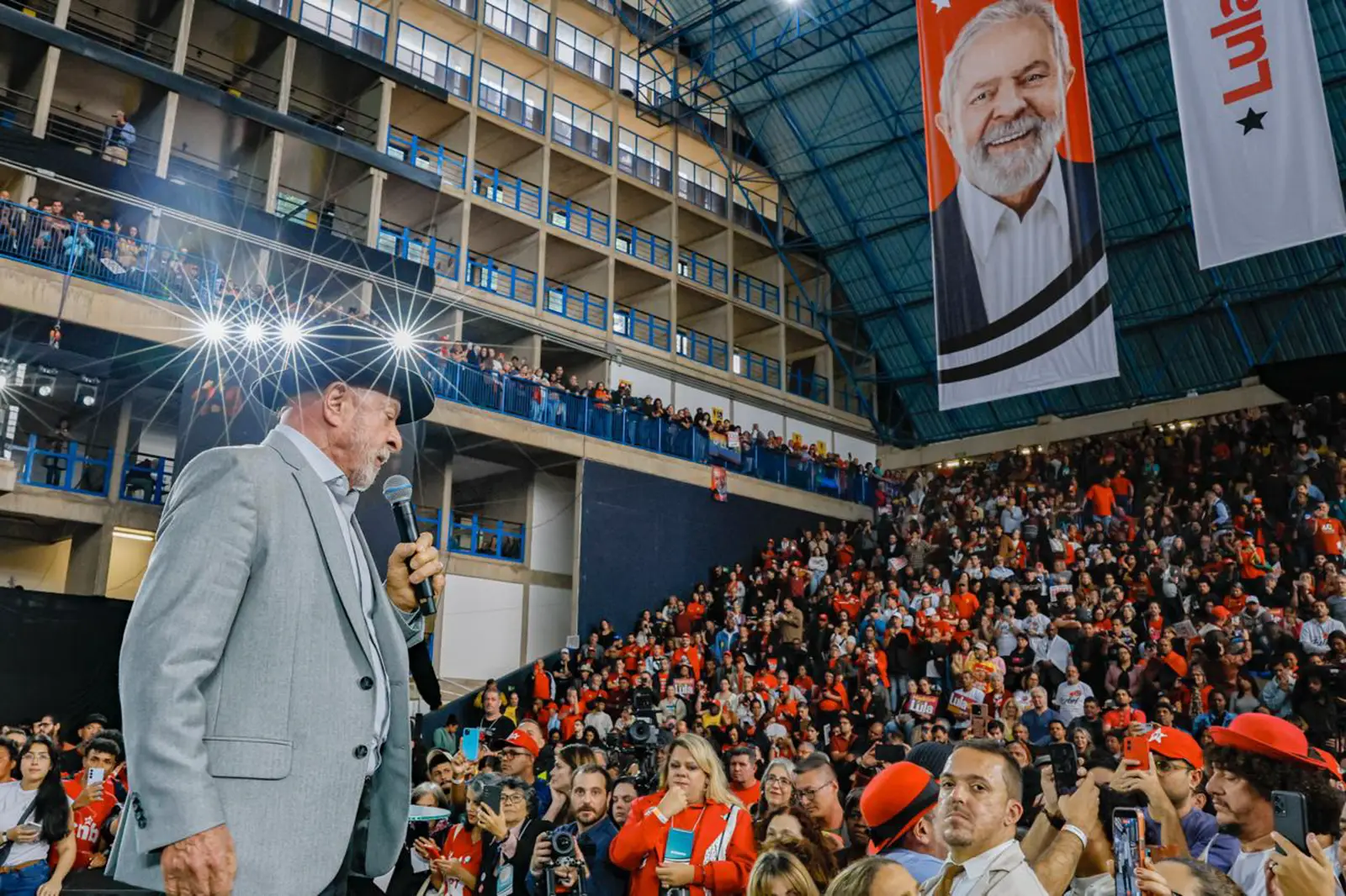 Luiz Inácio Lula da Silva, de perfil y con sombrero oscuro, habla con un micrófono en la mano ante un pabellón abarrotado de simpatizantes vestidos de rojo; al fondo cuelga una lona con su retrato