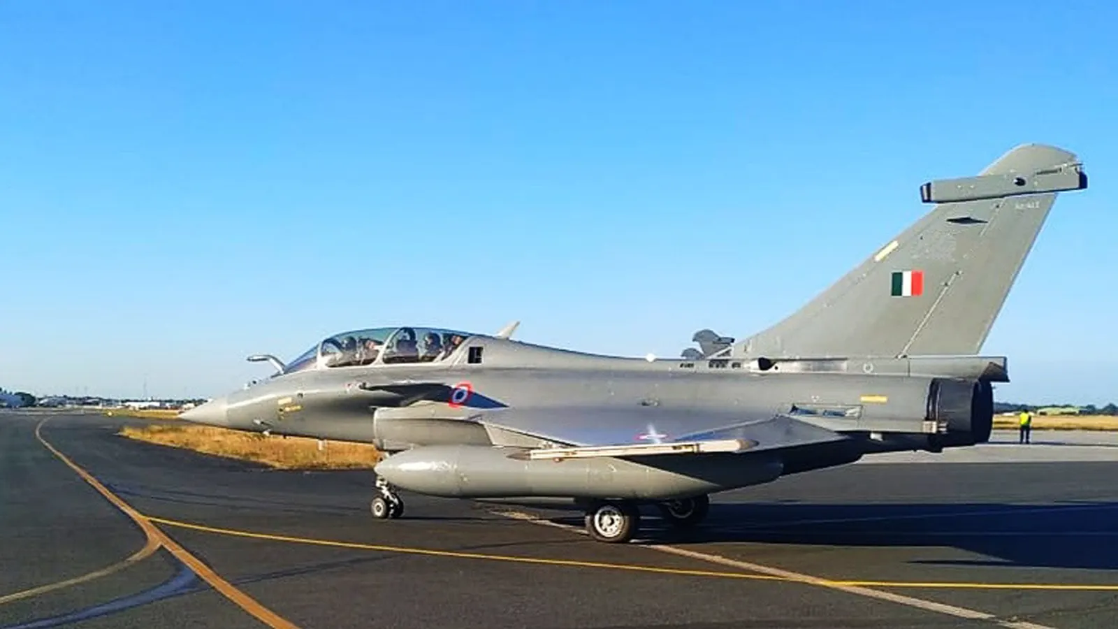 Un caza Rafale gris, con la bandera de India en el estabilizador vertical y dos tripulantes en la cabina, rueda por la pista de la factoría de Dassault Aviation en Mérignac bajo un cielo azul despejado.