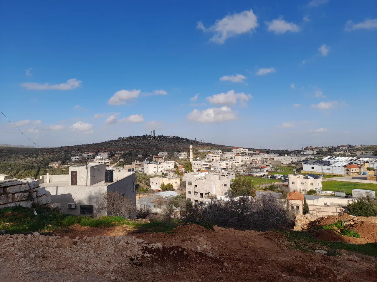 Vista general del pueblo palestino de Qusra, con casas blancas y el alminar de una mezquita, y al fondo una colina con el asentamiento israelí de Migdalim