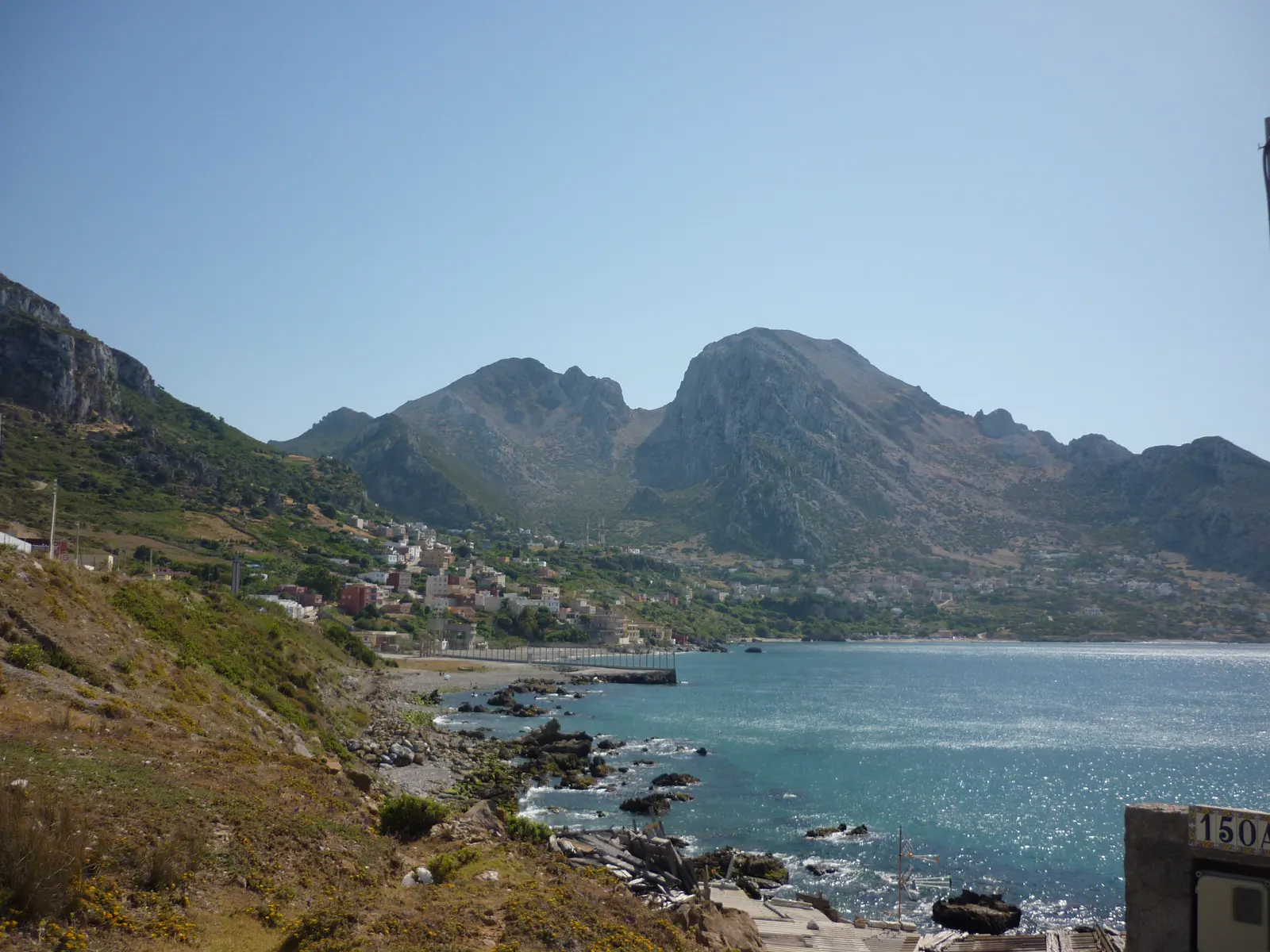 Vista de la bahía entre Benzú, en Ceuta, y la localidad marroquí de Beliones: una ladera de monte bajo en primer término, el mar a la derecha y, al fondo, el caserío marroquí al pie del monte Musa; la valla fronteriza baja por la orilla hasta el agua