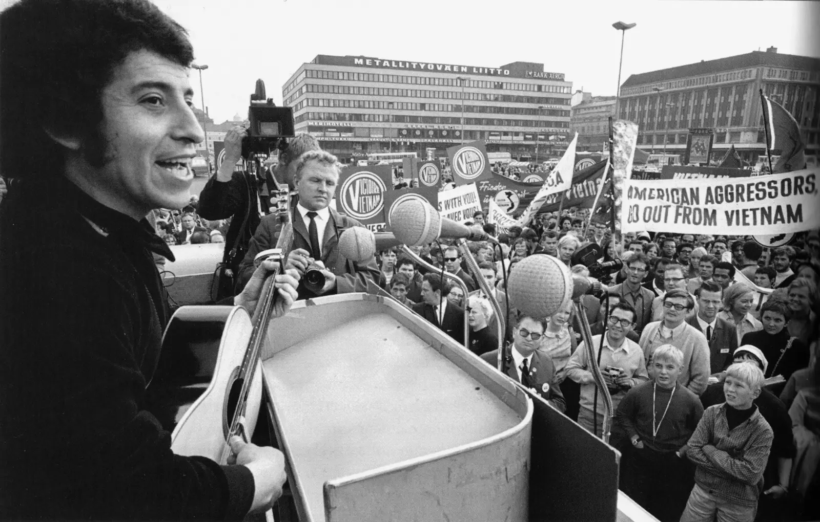 Fotografía en blanco y negro de Víctor Jara, con guitarra, cantando ante varios micrófonos en una plaza de Helsinki llena de gente que sostiene pancartas contra la guerra de Vietnam.