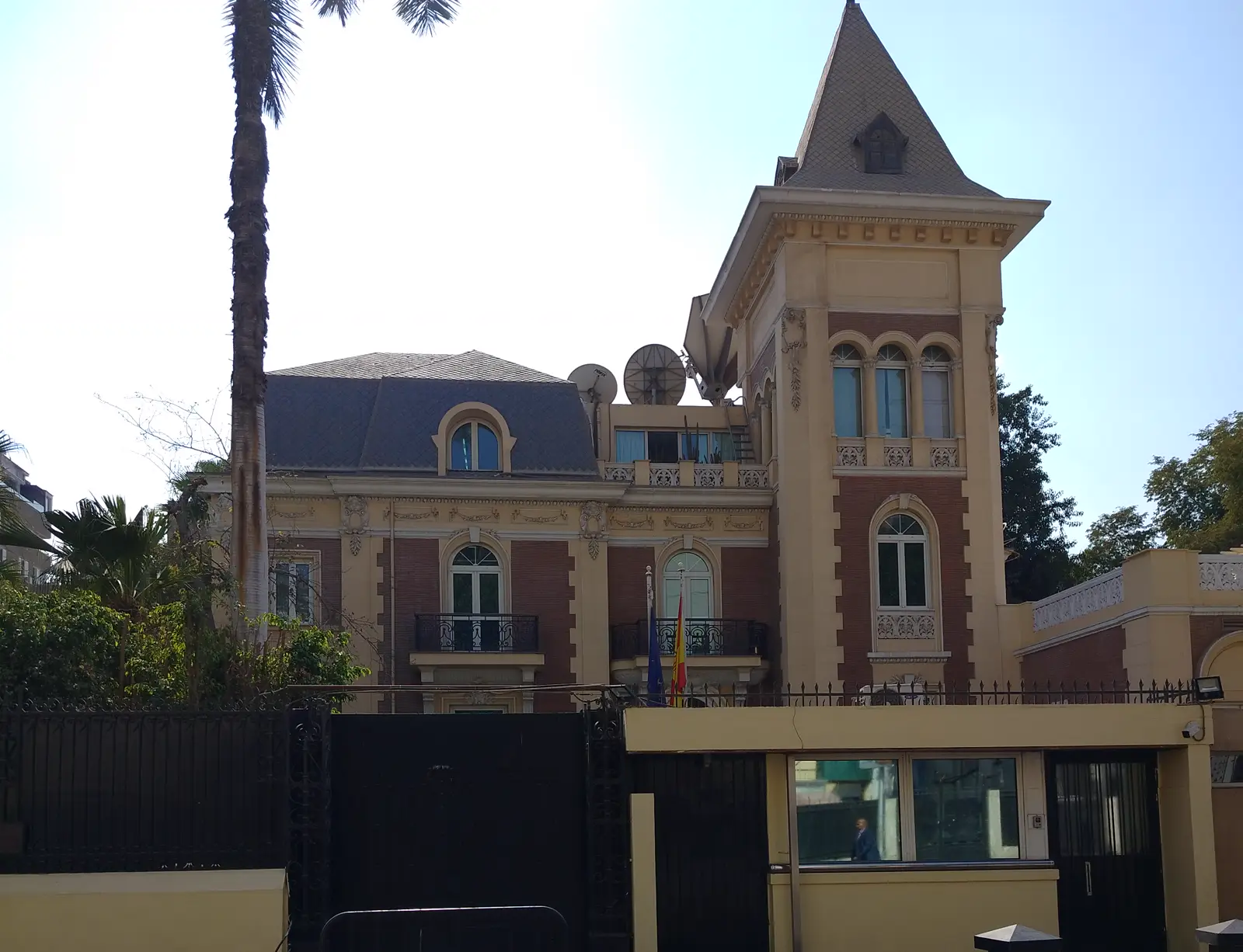 Fachada de la Embajada de España en El Cairo, un edificio de ladrillo y piedra con torreón y tejado de pizarra, con las banderas de España y de la Unión Europea en un balcón y una garita de seguridad ante la verja.