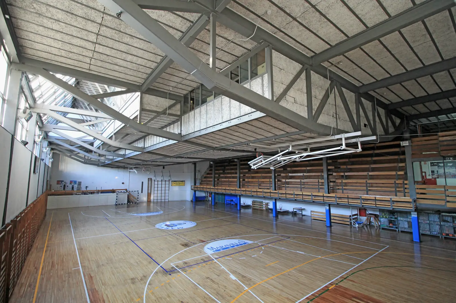 Interior vacío de un gimnasio escolar cubierto, con suelo de madera, líneas de cancha, gradas laterales y canasta plegada colgando de la estructura metálica del techo
