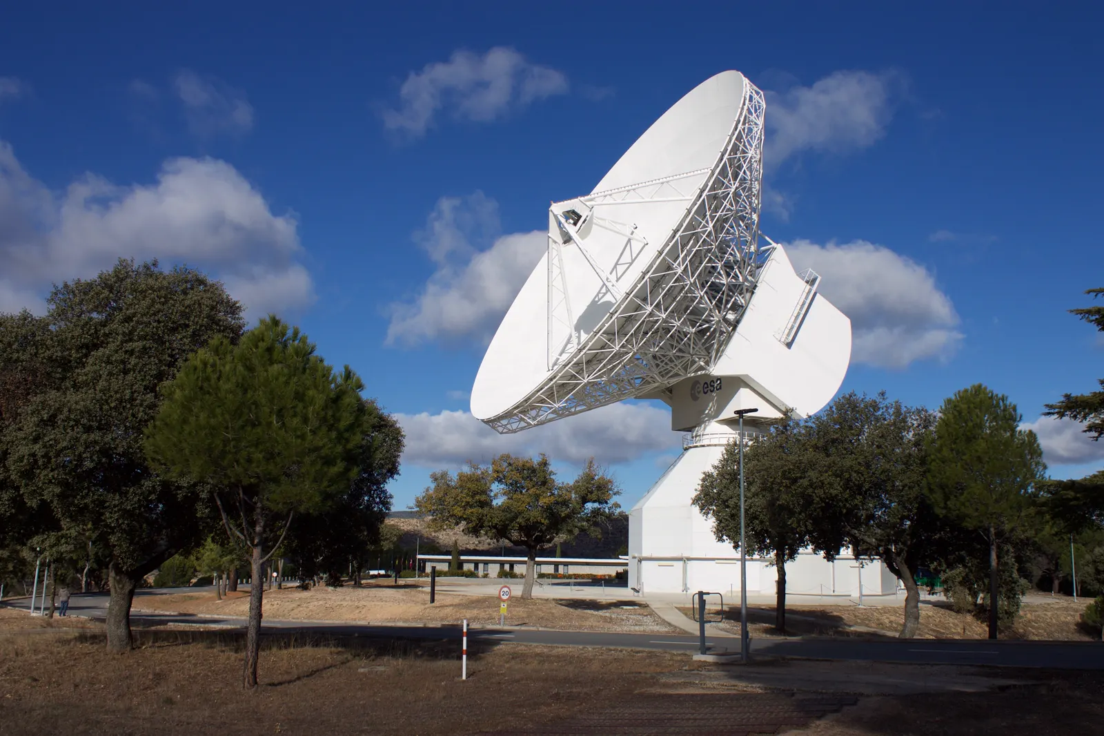 Gran antena parabólica blanca inclinada hacia el cielo sobre un pedestal cilíndrico, rodeada de encinas, pinos y pasto seco, con nubes al fondo.
