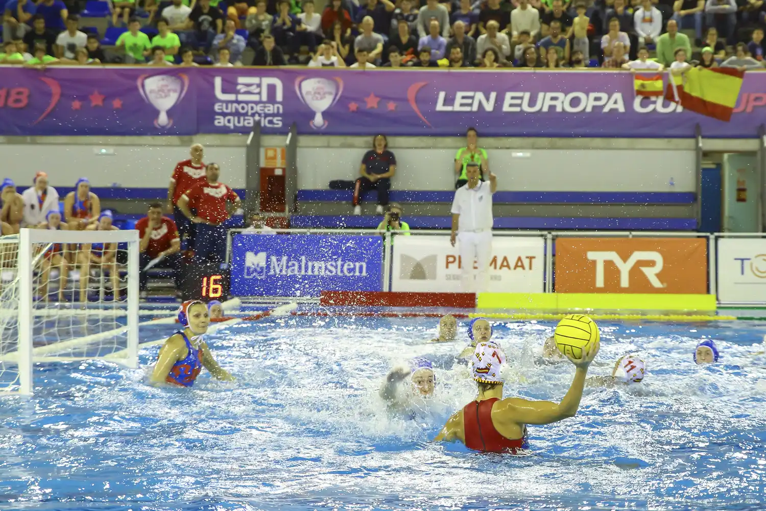 Una jugadora de la selección española de waterpolo, con bañador rojo y gorro blanco con el escudo de España, arma el brazo para lanzar el balón mientras dos rivales con gorro azul la marcan; al fondo, la portería, la portera y una lona de la LEN Europa Cup