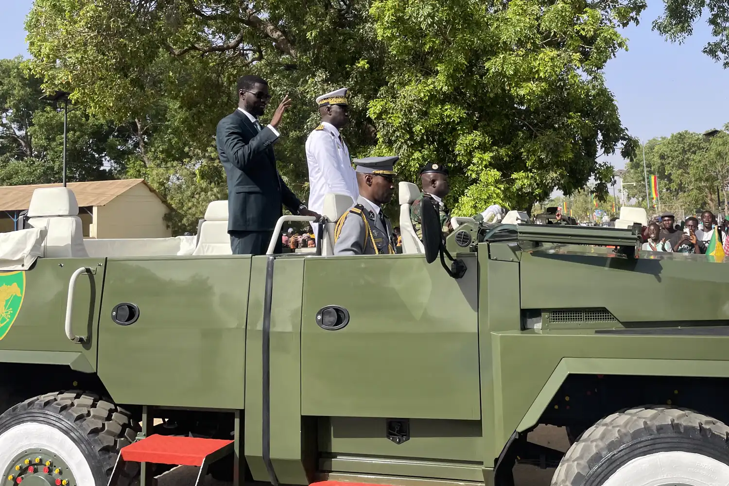 Un hombre trajeado saluda con la mano en alto de pie sobre un vehículo militar descubierto de color verde, acompañado por tres militares de uniforme, ante un público que le observa desde la acera