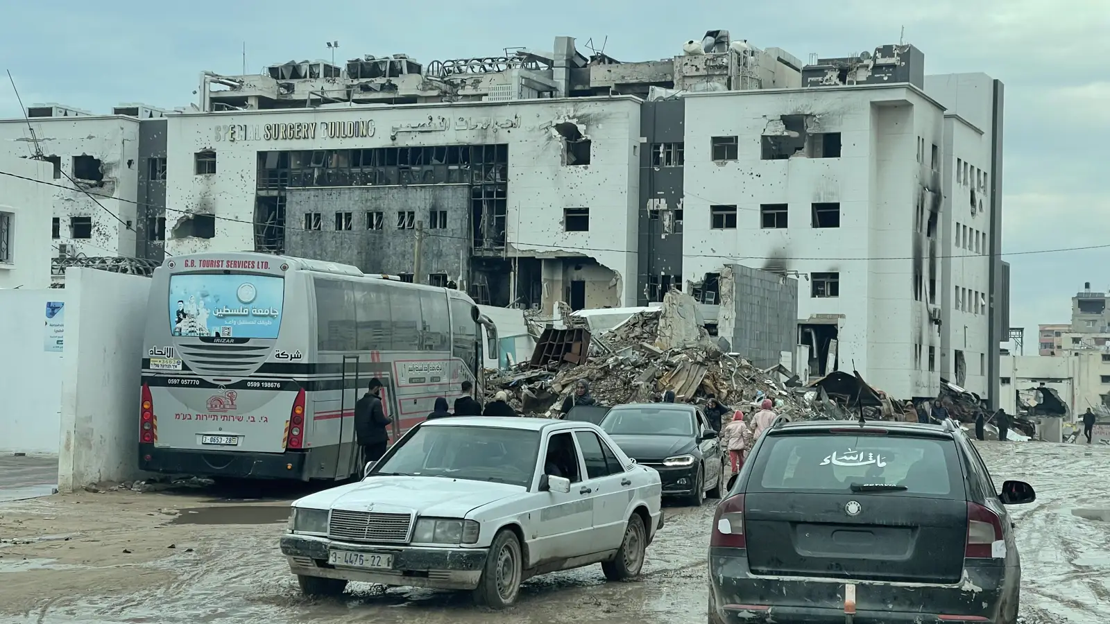 Fachada acribillada y calcinada del edificio de cirugía del hospital Al Shifa de Gaza, con escombros amontonados delante y coches y un autobús aparcados en la explanada