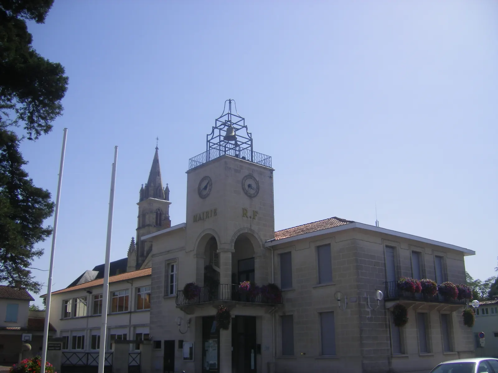 Fachada del ayuntamiento de Marcheprime, un edificio de piedra clara con una torre de reloj rematada por una campana bajo una estructura metálica, con macetas de flores en los balcones y el campanario de la iglesia asomando detrás, bajo un cielo despejado.