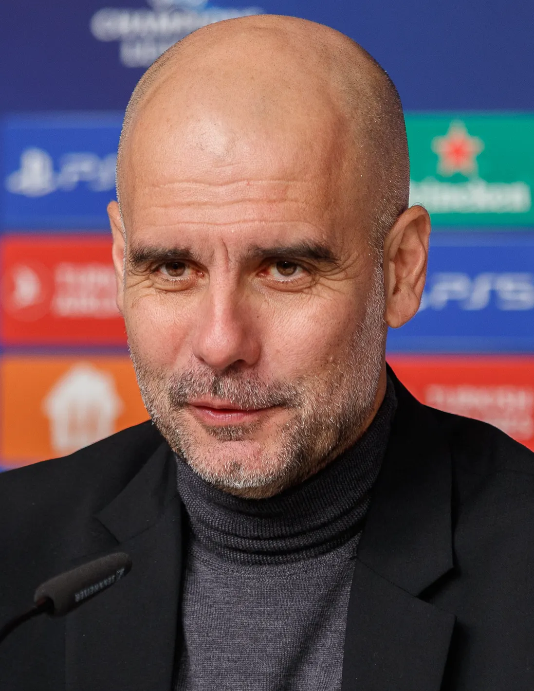 Primer plano de Pep Guardiola, con jersey gris de cuello alto y chaqueta negra, ante un micrófono y un panel de patrocinadores de la Liga de Campeones