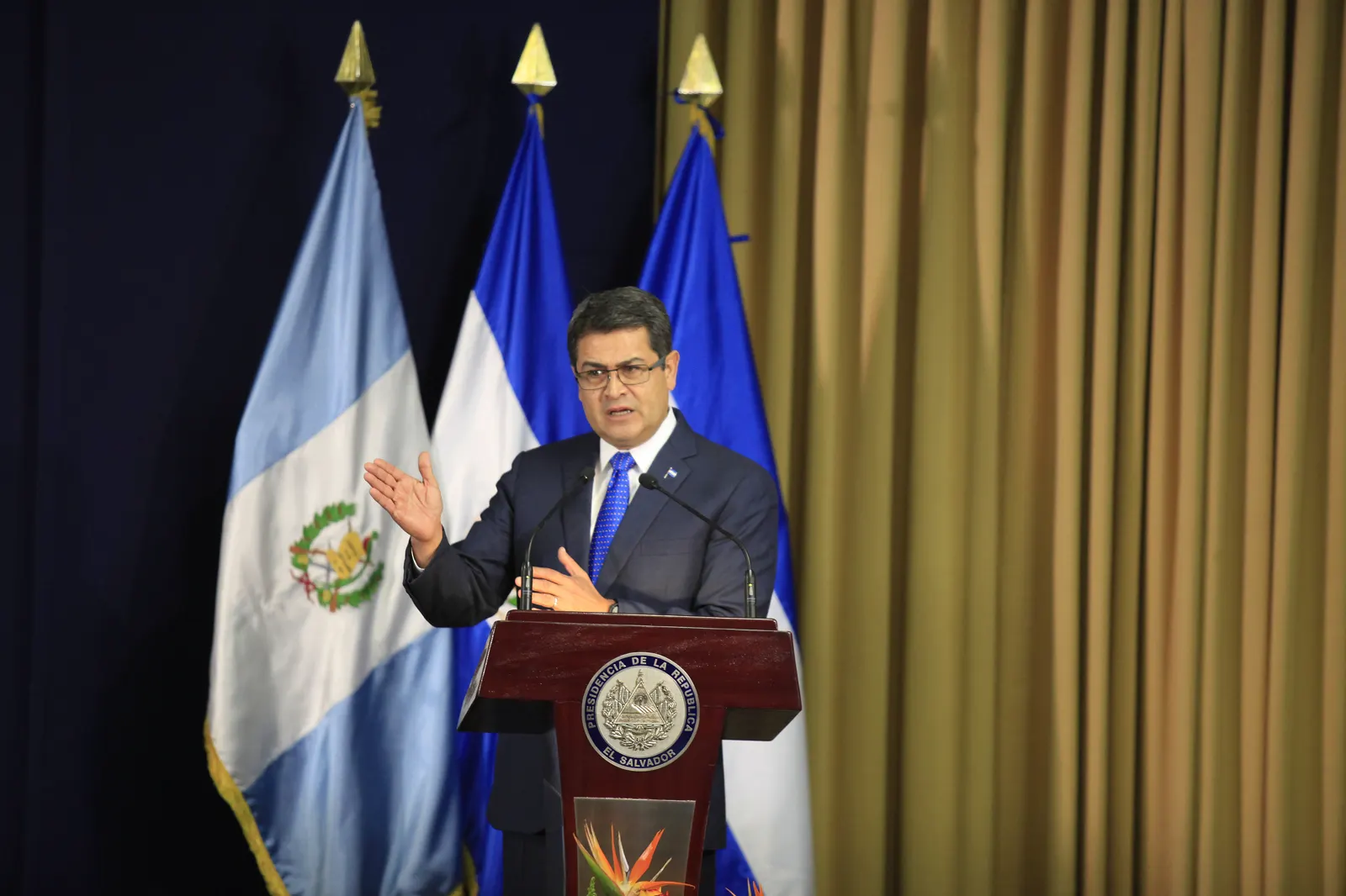 Juan Orlando Hernández, con traje oscuro y gafas, habla en un atril con el escudo de la Presidencia de El Salvador, ante banderas de Guatemala y El Salvador.