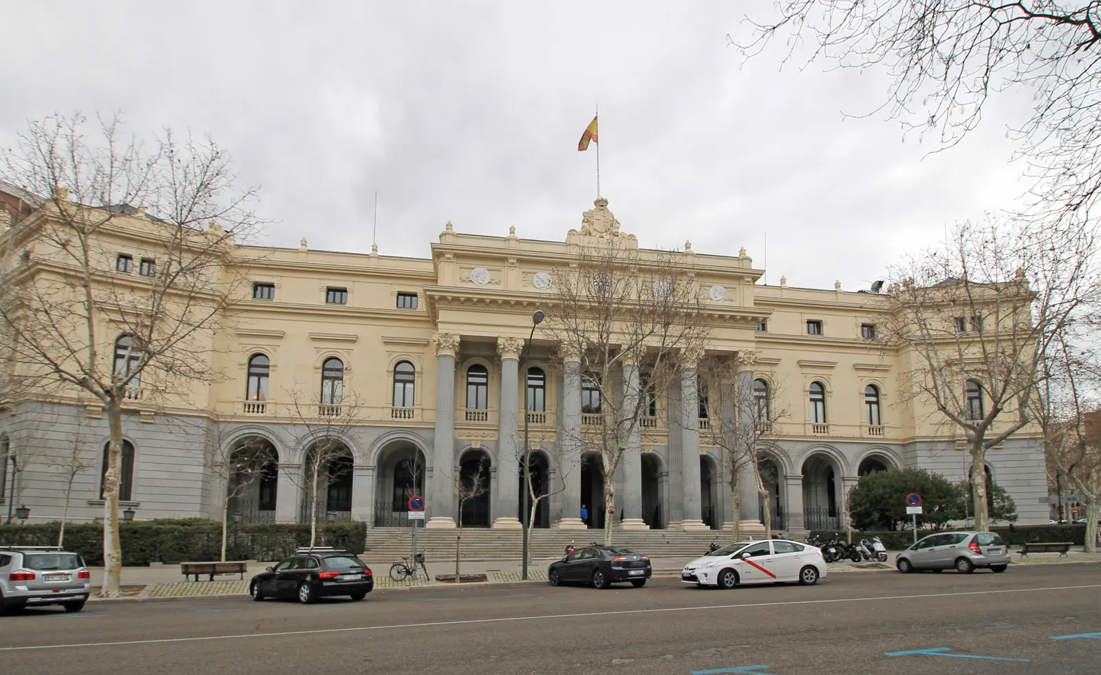 Fachada del Palacio de la Bolsa de Madrid, edificio neoclásico de piedra clara con columnas y bandera de España en la cubierta, con árboles sin hojas y coches aparcados delante.