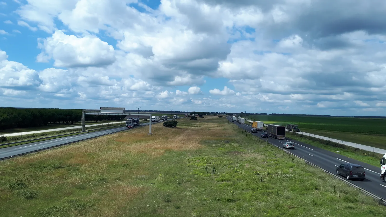Vista elevada de la autopista A63 a su paso por Cestas, en Gironda: dos calzadas separadas por una amplia mediana de hierba, con camiones y turismos circulando en ambos sentidos, campos verdes a los lados y cielo con nubes.
