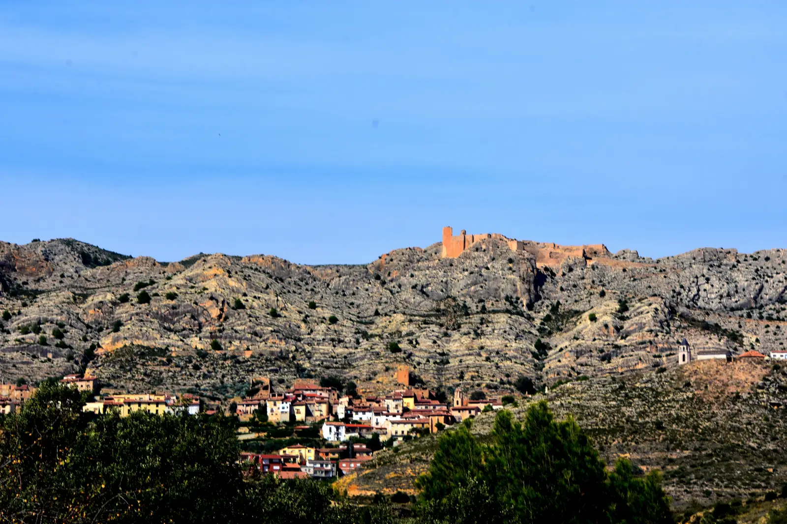 Vista general del pueblo de Castellote (Teruel), con casas de tejado rojo al pie de una ladera rocosa coronada por las ruinas de su castillo.