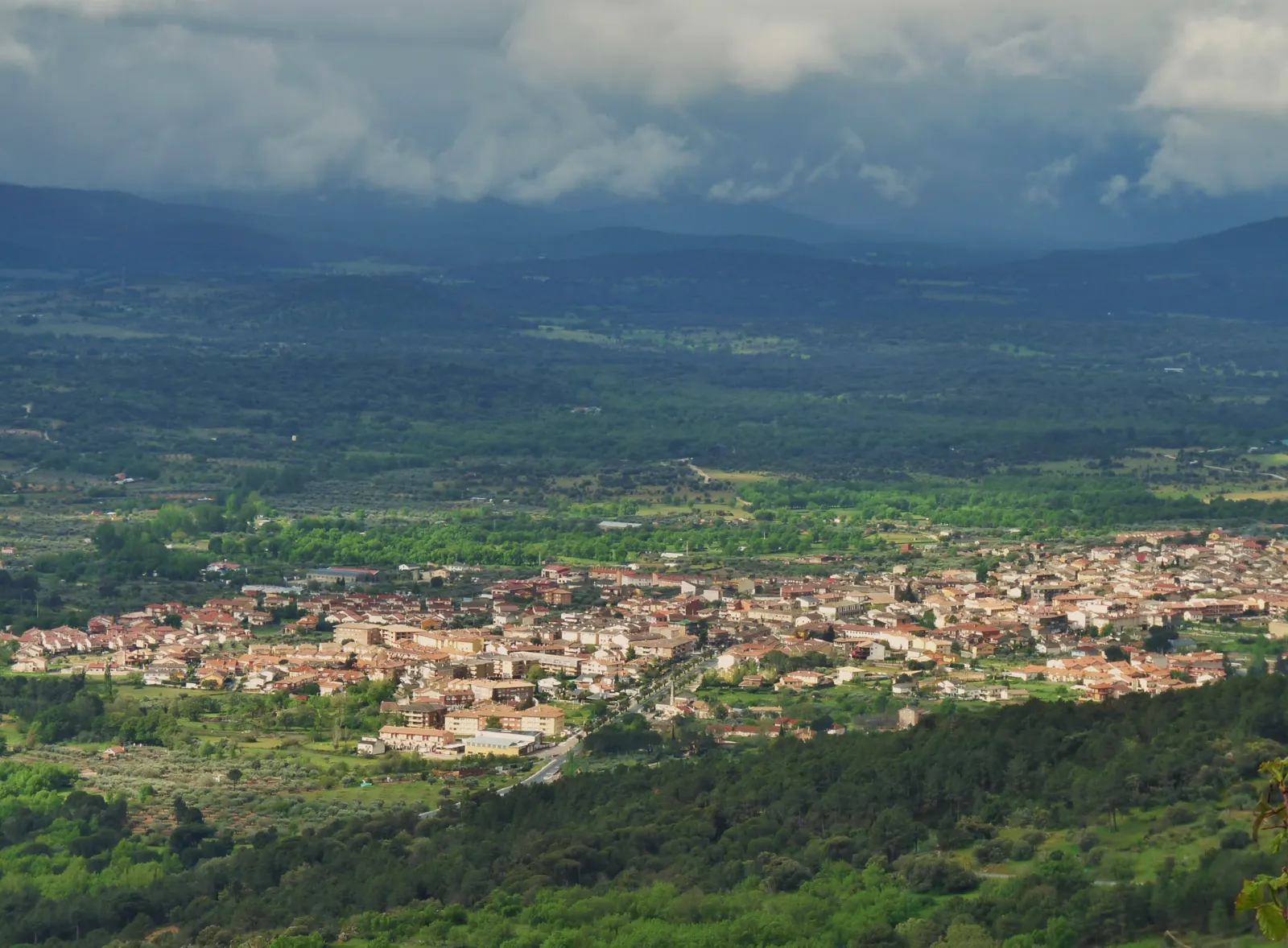 Vista aérea de Sotillo de la Adrada (Ávila) desde una ladera: el casco urbano de tejados rojizos ocupa el fondo del valle, rodeado de olivares, pinares y monte bajo, con las sierras del Valle del Tiétar y nubes bajas al fondo.