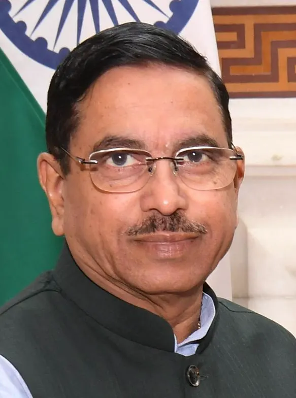 Retrato de Pralhad Joshi, hombre de mediana edad con gafas, bigote y chaqueta oscura sin cuello, ante la bandera de India.
