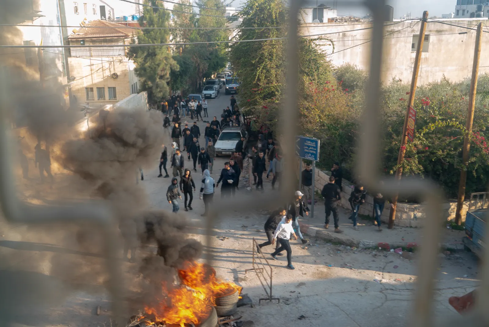 Vista desde una ventana de una calle del campo de refugiados de Yenín durante una operación militar israelí: decenas de jóvenes palestinos caminan entre edificios de hormigón mientras arde un montón de neumáticos en primer plano
