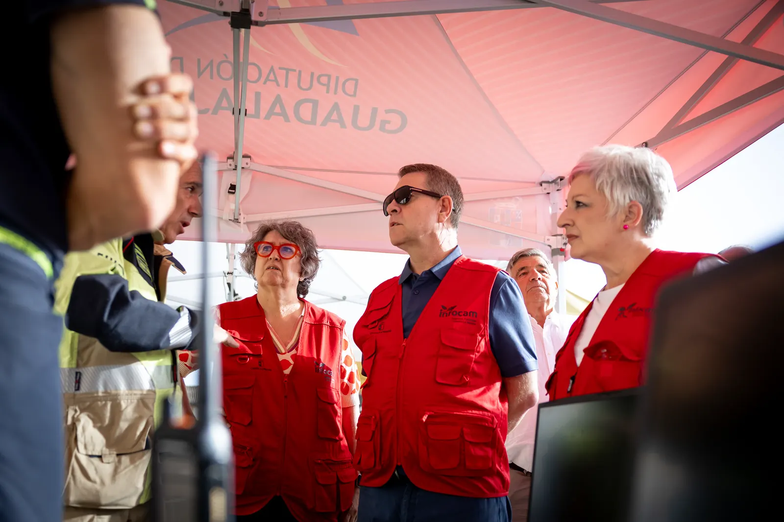 Bajo una carpa blanca y roja con el rótulo «Diputación de Guadalajara», cuatro personas con chalecos rojos del dispositivo Infocam escuchan a un hombre de uniforme con banda reflectante que señala hacia un ordenador portátil.