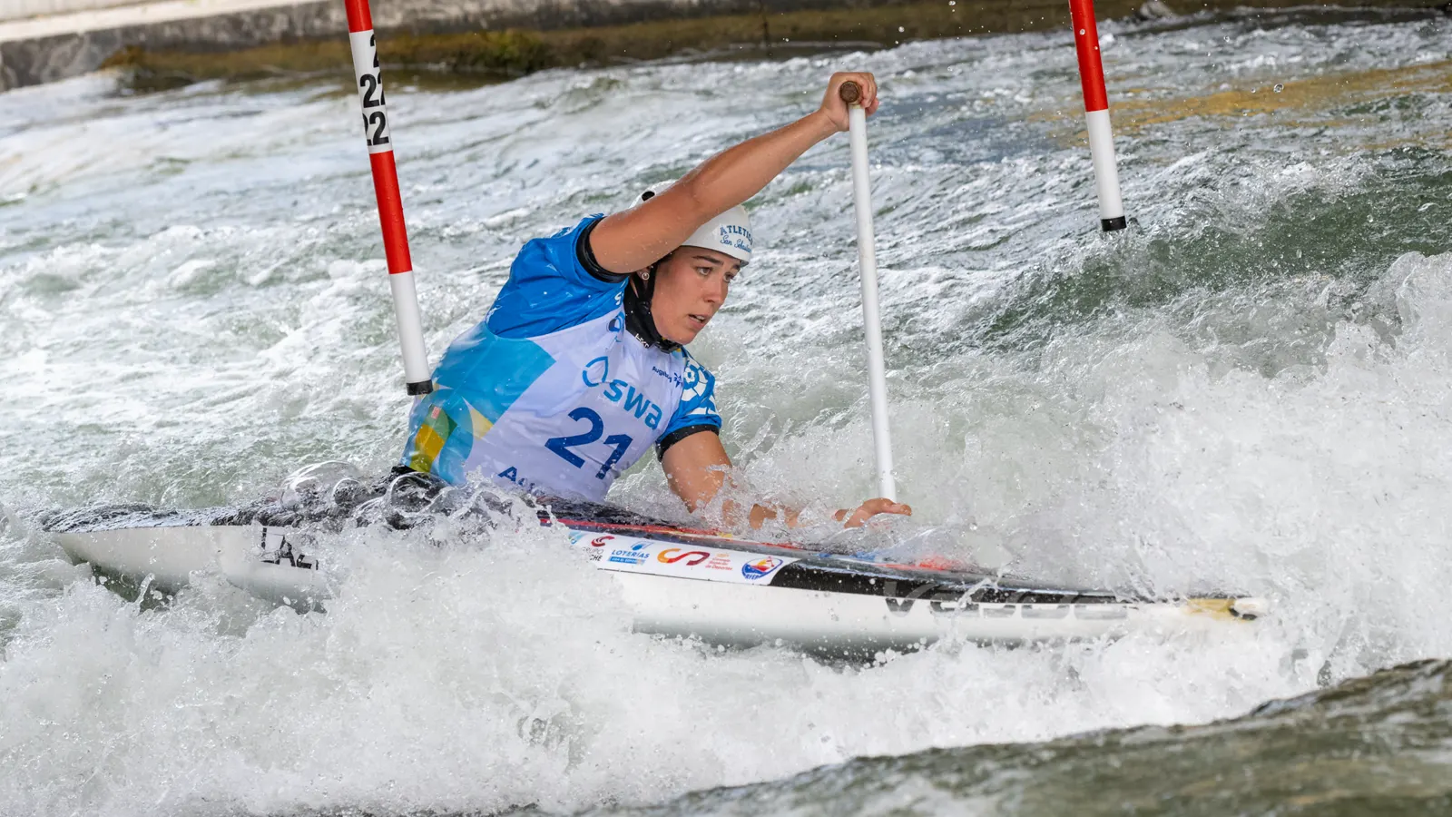 La piragüista Miren Lazkano, con casco blanco y peto azul con el dorsal 21, rema arrodillada en una canoa entre la espuma de un canal de aguas bravas, junto a dos palos rojiblancos de una puerta de eslalon.