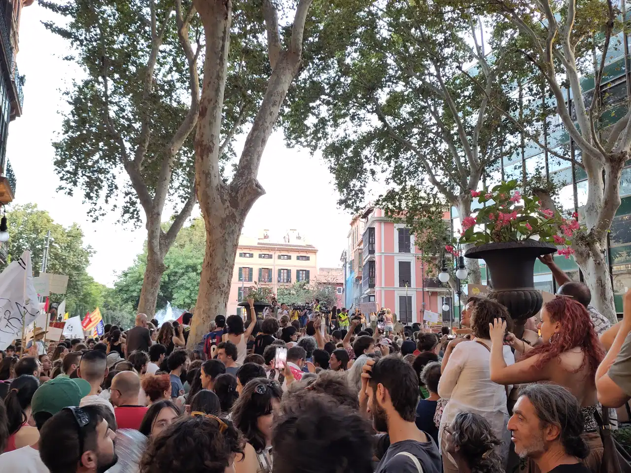 Multitud compacta de manifestantes bajo los plátanos de un paseo de Palma; al fondo, entre fachadas de colores, un escenario con altavoces y varias personas de pie ante la gente.