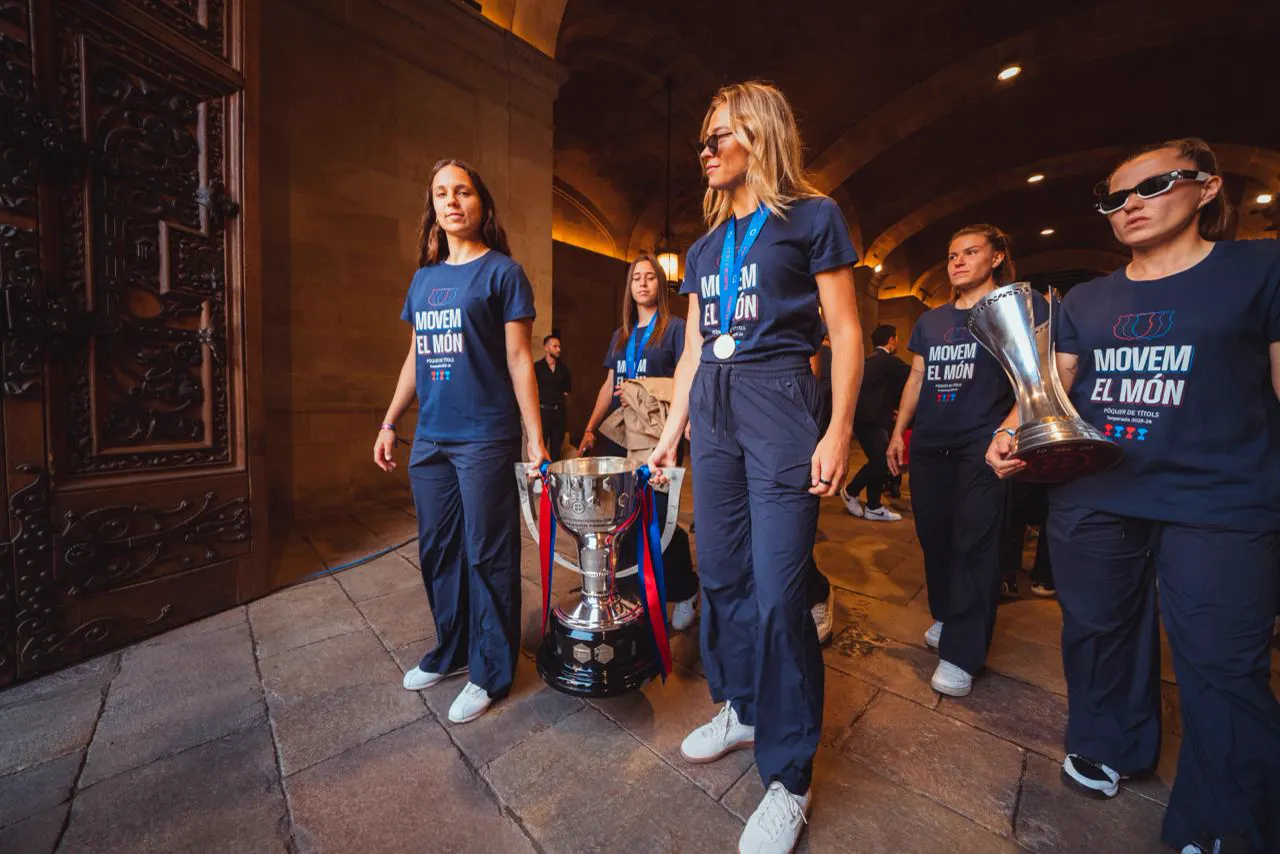 Cinco jugadoras del Barça Femenino, con camiseta azul marino que reza «Movem el món», caminan bajo los arcos de piedra del Palau de la Generalitat llevando dos trofeos plateados