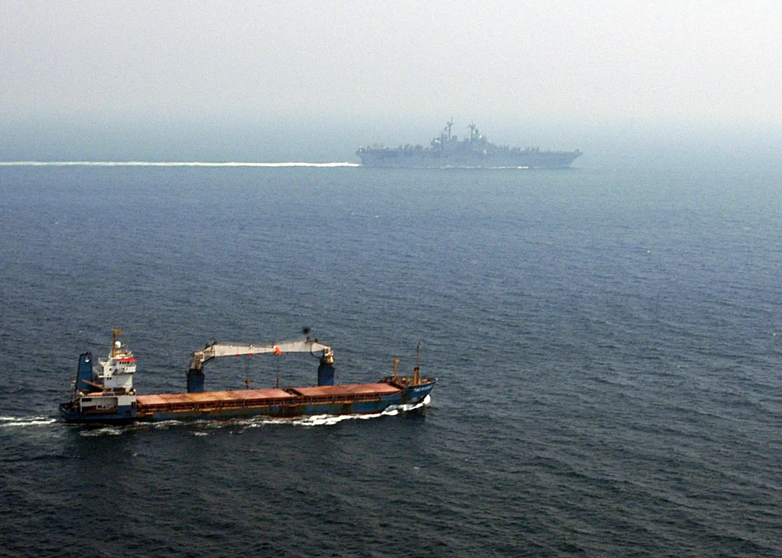 Vista aérea del mar: en primer plano, un carguero azul con grúas de cubierta navega por el estrecho; al fondo, entre la calima, un gran buque de guerra estadounidense.