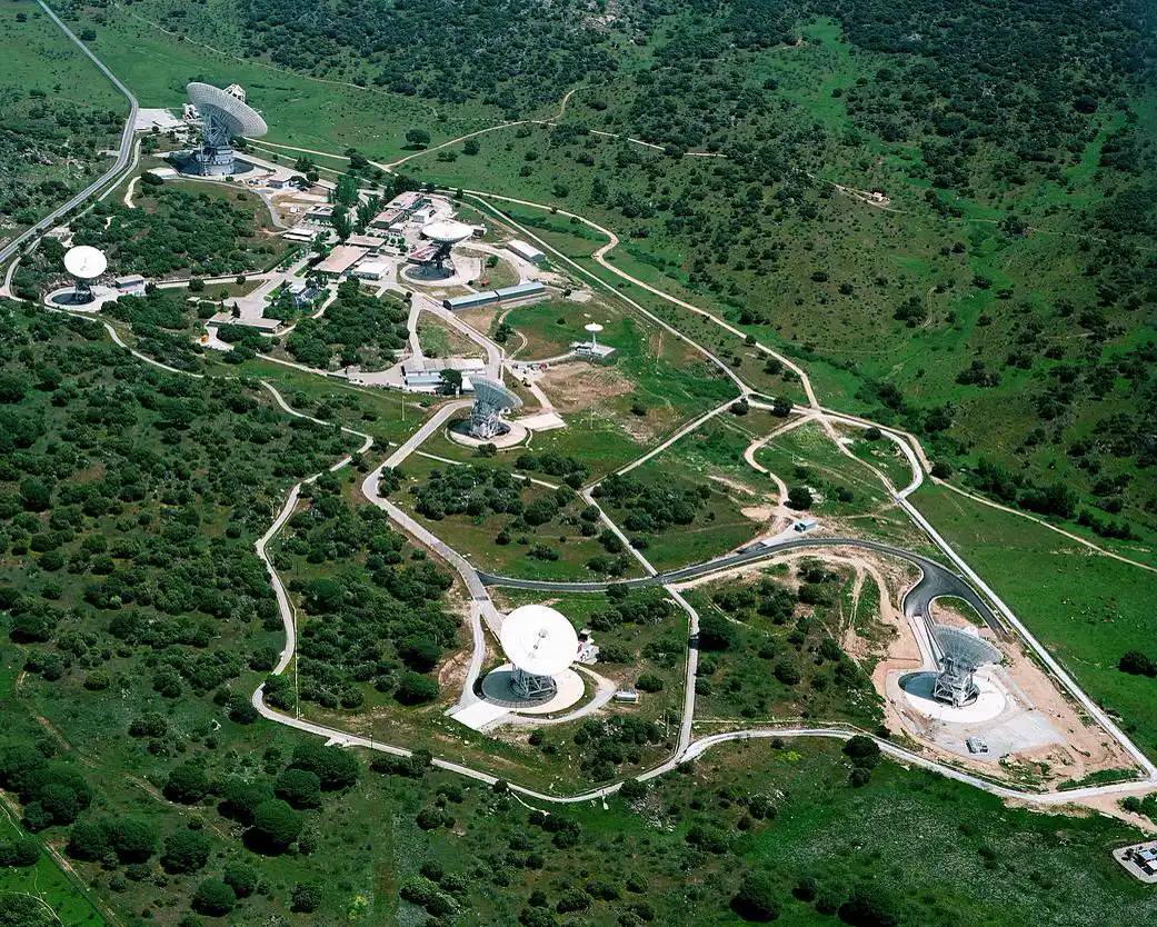 Vista aérea del complejo de antenas de Robledo de Chavela: seis grandes parábolas blancas repartidas por una loma de monte bajo, unidas por carreteras y con varios edificios de una planta en el centro.