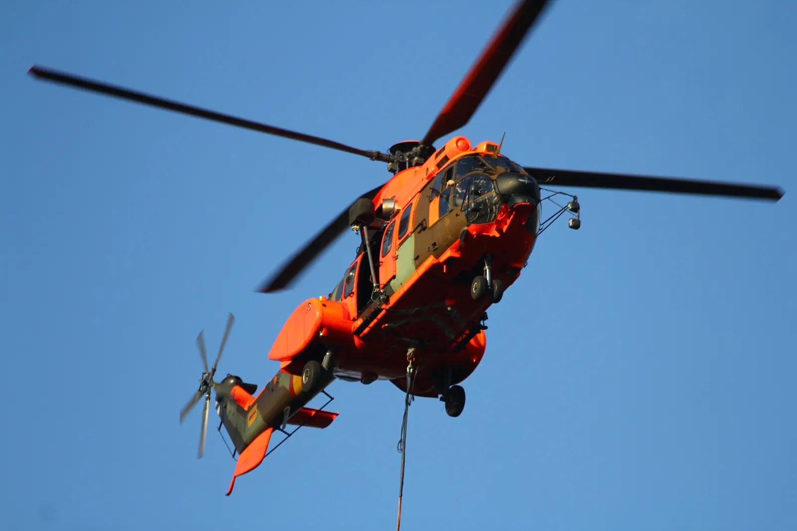 Helicóptero Cougar naranja y verde del Ejército de Tierra en vuelo contra un cielo azul, con un cable de carga colgando, durante labores de extinción.