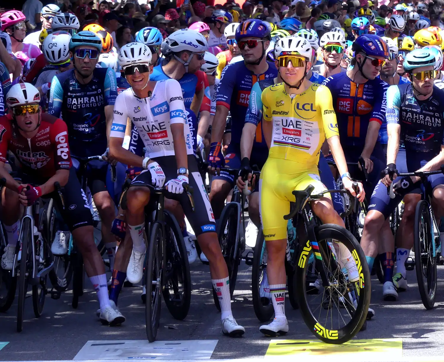 Tadej Pogacar, con el maillot amarillo del UAE Team Emirates-XRG, y su compañero Isaac del Toro, con el maillot blanco, esperan sonrientes sobre sus bicicletas en la línea de salida de una etapa, rodeados de corredores de otros equipos.