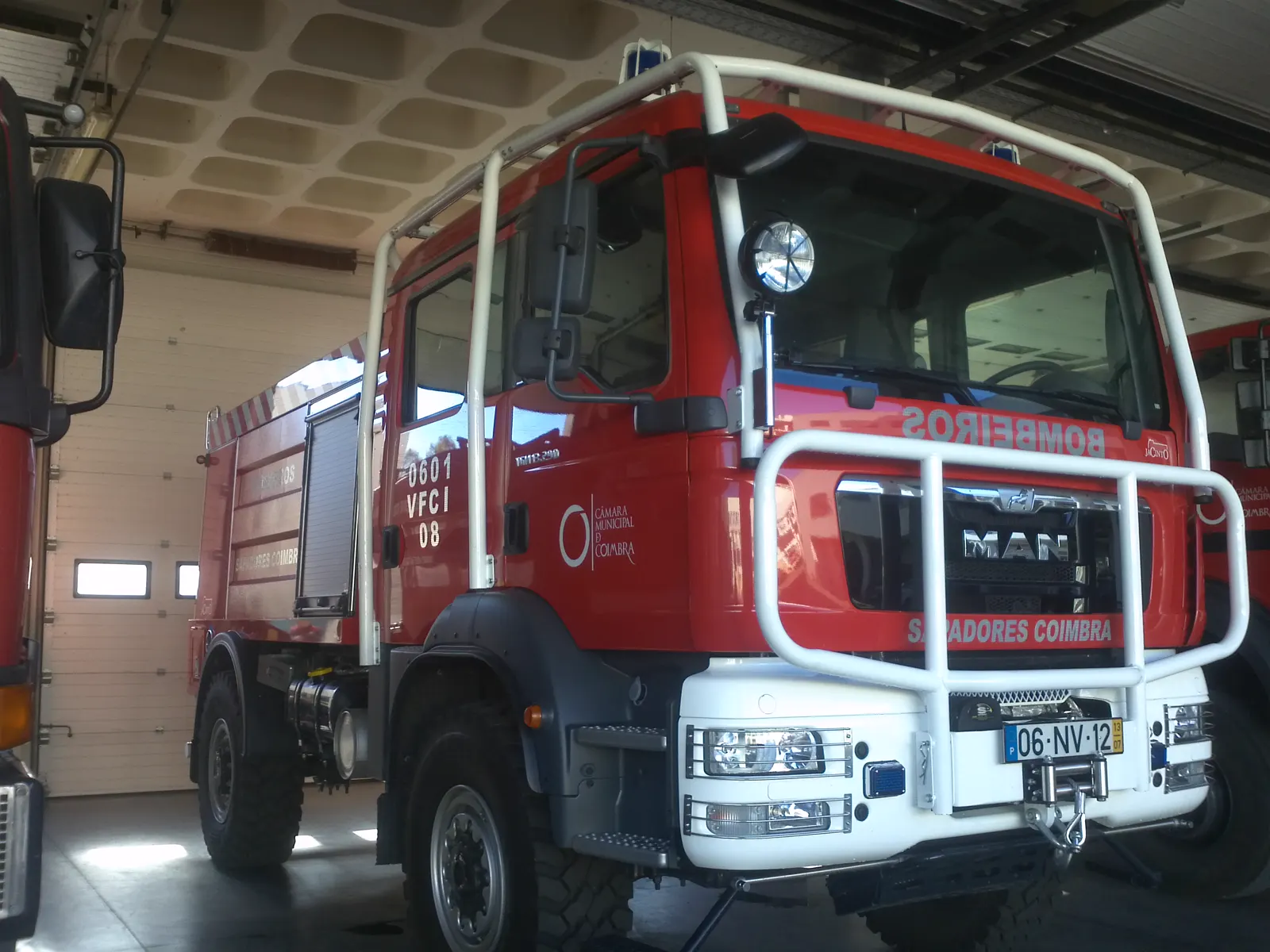 Camión rojo de combate a incendios forestales de los Bomberos Sapadores de Coímbra, con matrícula portuguesa y defensa delantera blanca, aparcado en el interior de un parque de bomberos.