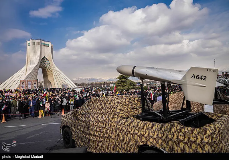 Dron Shahed-136 de fuselaje blanco y ala en delta, con el código A642 pintado en la deriva, montado sobre una rampa de lanzamiento en la caja de una furgoneta cubierta con red de camuflaje; al fondo, la torre Azadi de Teherán y una multitud con banderas iraníes.