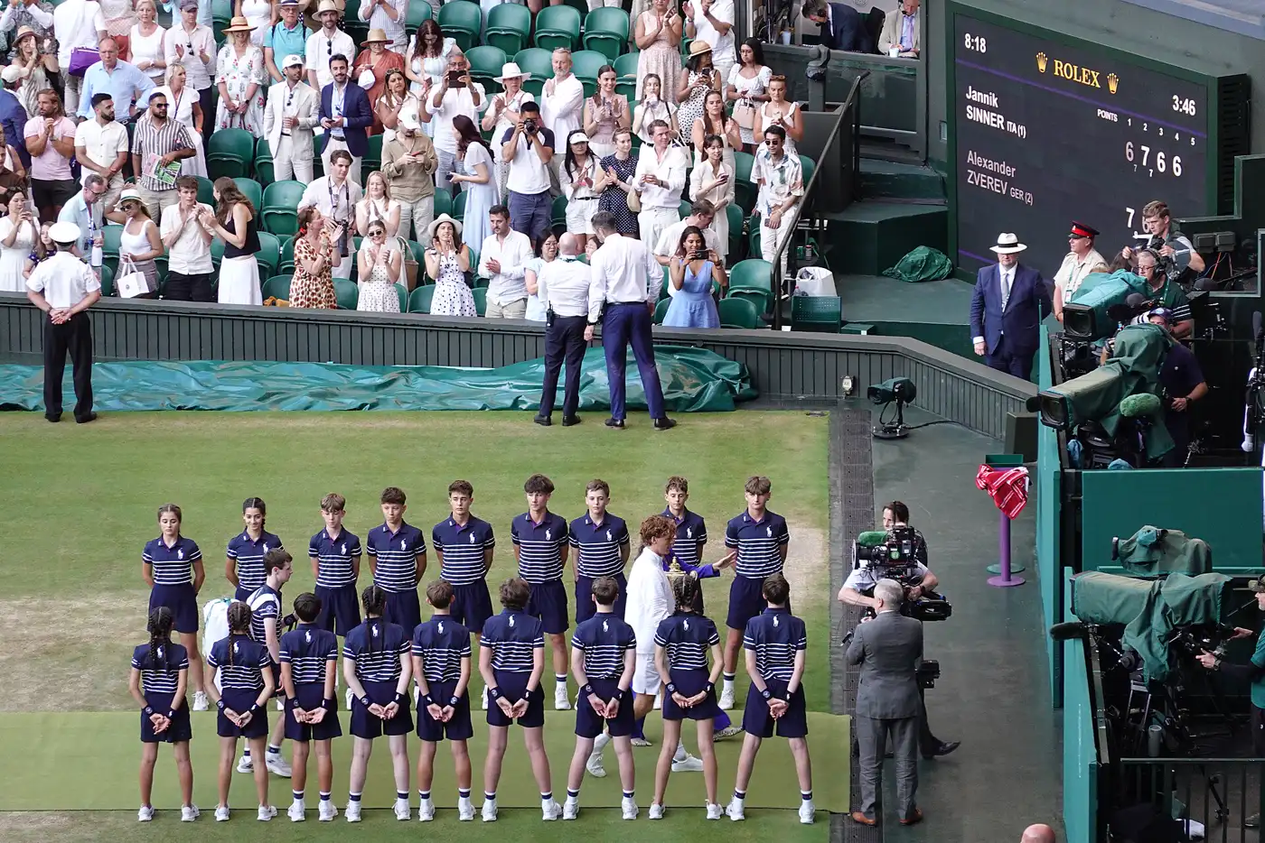 Jannik Sinner, vestido de blanco, sostiene un trofeo dorado en el césped de la pista central de Wimbledon, rodeado por dos filas de recogepelotas con uniforme azul marino; al fondo, el público en pie y el marcador con su nombre y el de Alexander Zverev