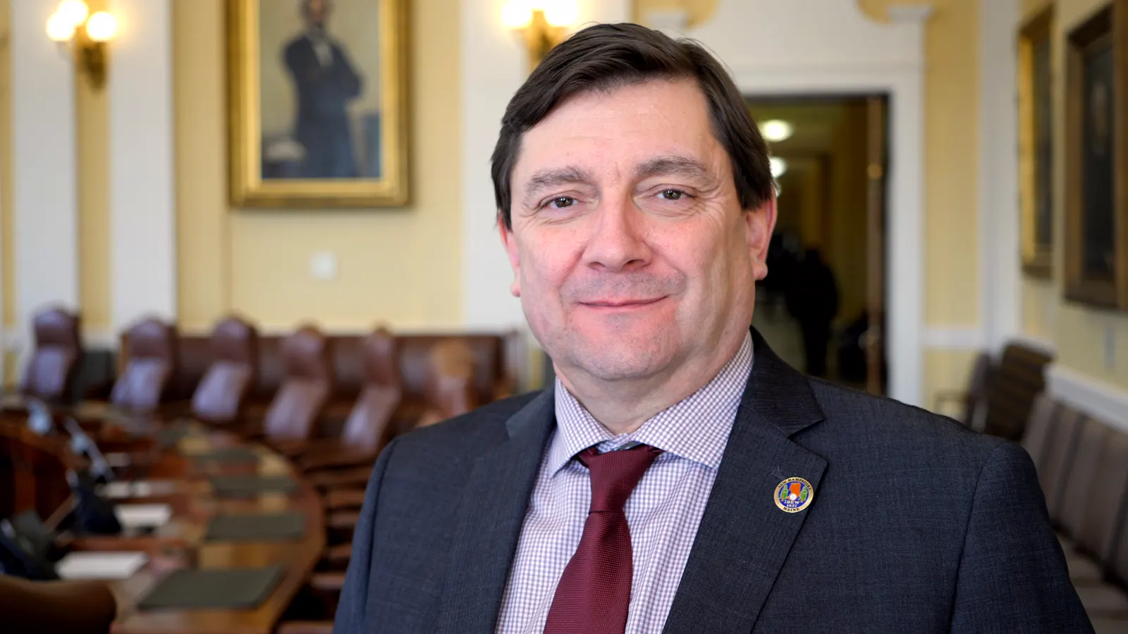 Retrato de Troy Jackson, con traje gris y corbata granate, de pie ante los escaños de cuero del hemiciclo del Senado de Maine.