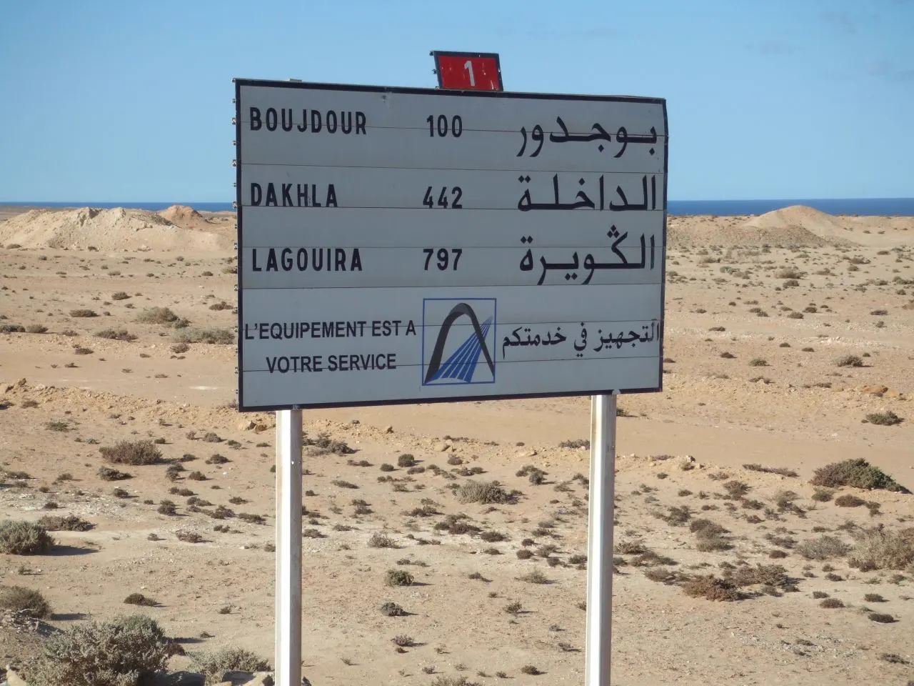 Señal de tráfico metálica al borde de una carretera del desierto, con las distancias en francés y árabe a Bojador (100), Dajla (442) y La Agüera (797); al fondo, llanura pedregosa y el océano Atlántico