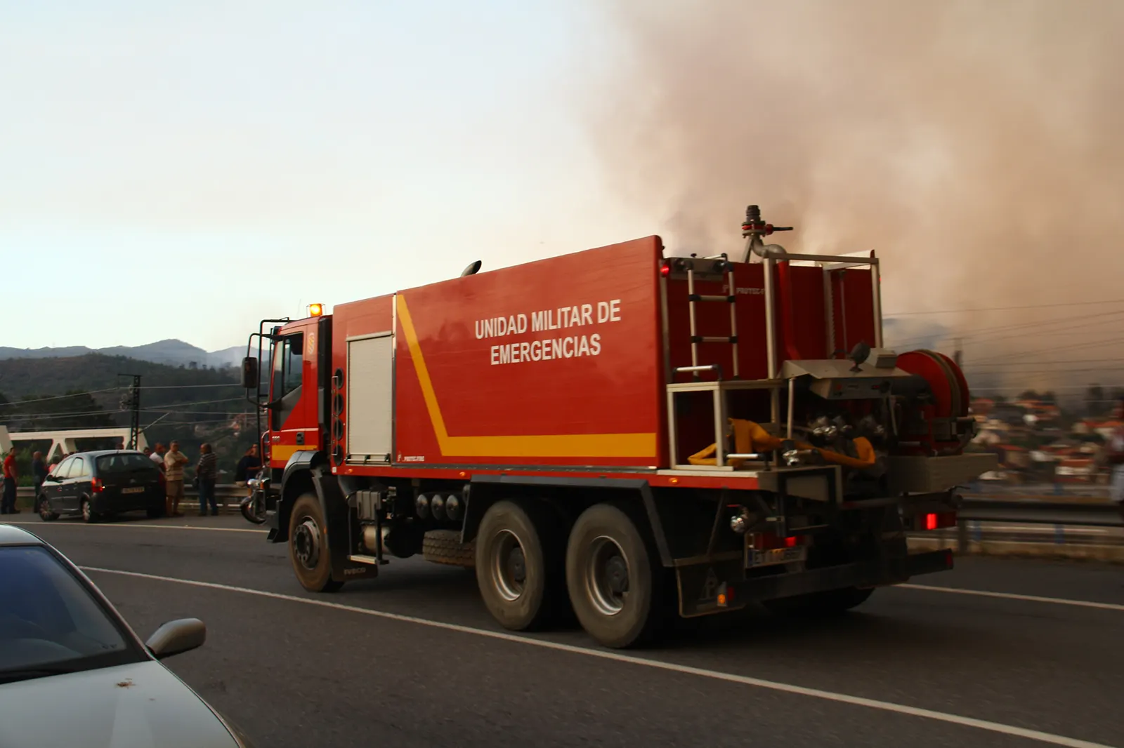 Camión autobomba rojo con el rótulo «Unidad Militar de Emergencias» circulando por una carretera hacia una gran columna de humo que cubre el cielo sobre un valle; a la izquierda, varios vecinos observan el incendio desde el arcén.
