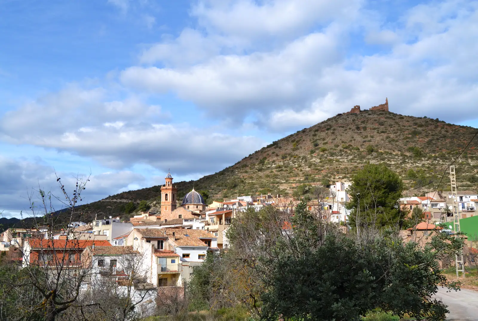 Casco urbano de Azuébar, con el campanario y la cúpula azul de la iglesia entre tejados de teja, al pie de un cerro de monte bajo coronado por las ruinas de un castillo.