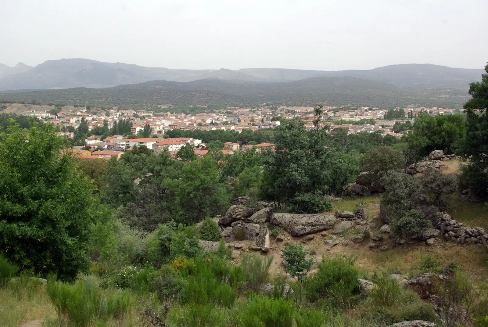 Vista de Navaluenga (Ávila) desde una ladera al sur: el casco urbano de tejados rojizos se extiende en el fondo del valle, rodeado de encinares y pinares, con bolos de granito y matorral en primer plano y la sierra al fondo.
