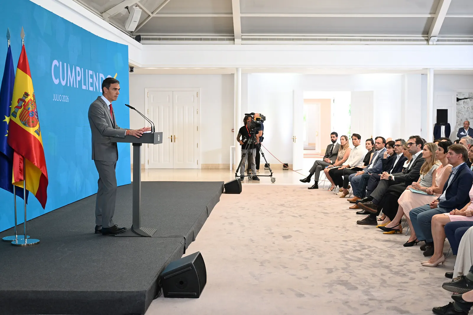 Pedro Sánchez, de traje gris, habla en un atril sobre una tarima en el Palacio de la Moncloa, con las banderas de España y de la Unión Europea a su izquierda y un panel azul con el lema «Cumpliendo. Julio 2026» al fondo; frente a él, una cámara de televisión y una fila de asistentes sentados.