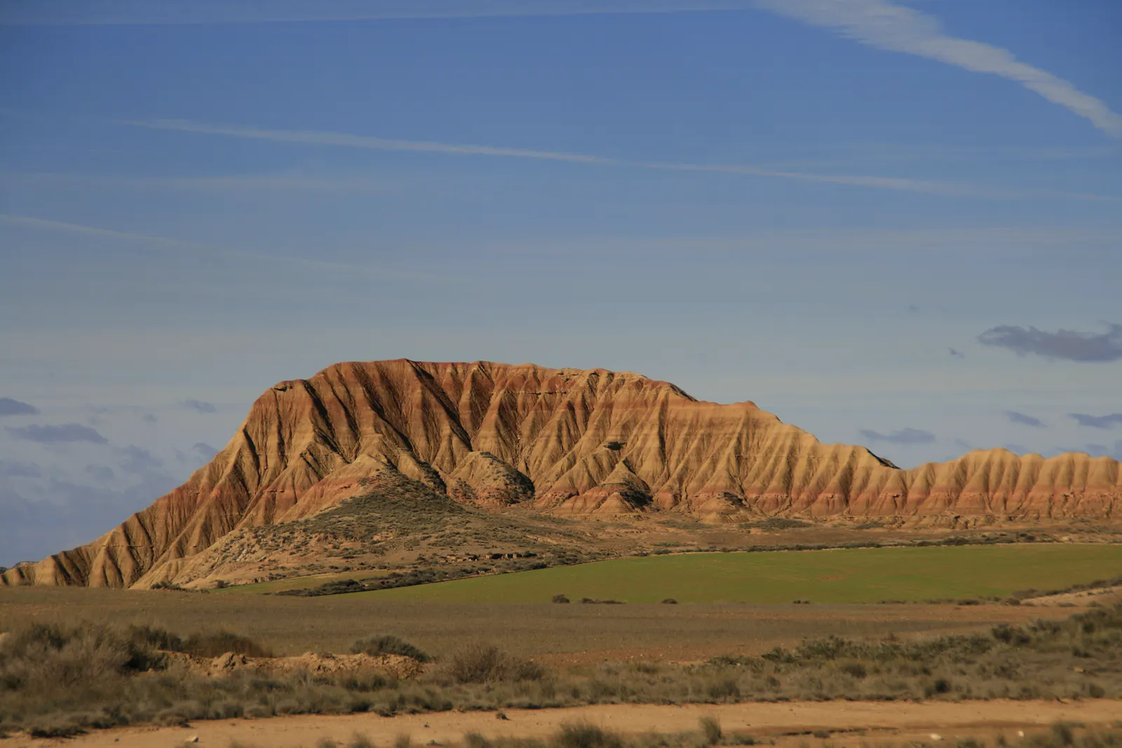 Paisaje semidesértico de las Bardenas Reales, en el sureste de Navarra: un cerro testigo de laderas erosionadas se levanta sobre un llano de tierra clara y matorral ralo, bajo un cielo despejado.