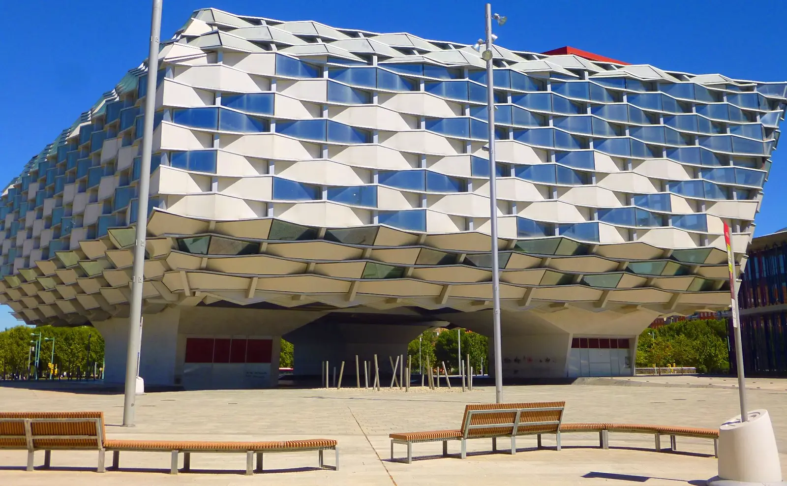 Fachada del Pabellón de Aragón, un edificio blanco de planta ovalada con celosía de lamas entrelazadas y ventanales, elevado sobre pilares, con una explanada de bancos vacíos en primer plano
