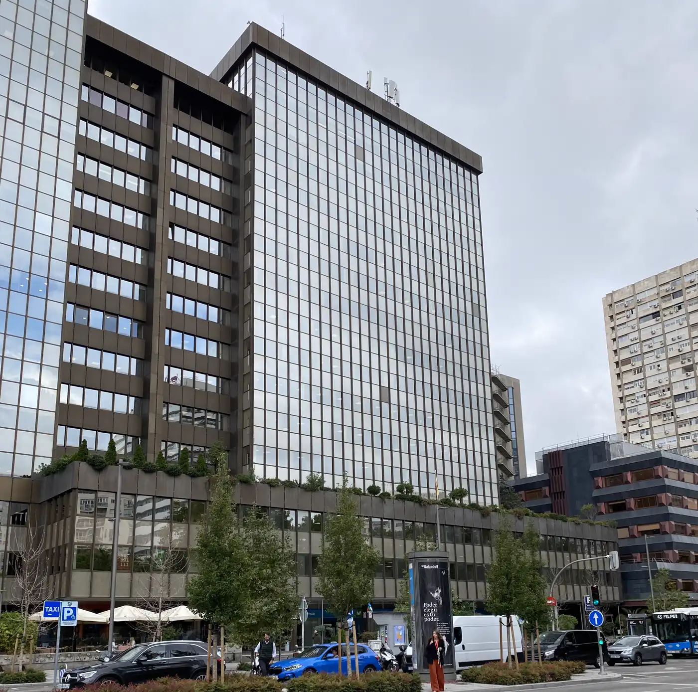 Torre de oficinas de fachada acristalada y perfilería oscura en el complejo AZCA de Madrid, vista desde la calle, con árboles, coches y viandantes en primer término bajo un cielo nublado.