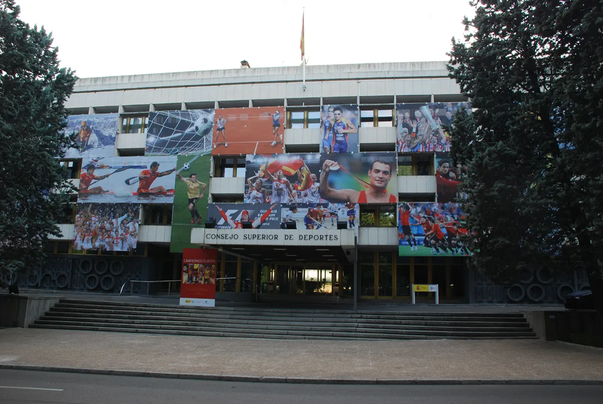 Fachada del edificio del Consejo Superior de Deportes en Madrid, con el rótulo «Consejo Superior de Deportes» sobre la entrada, grandes lonas fotográficas de deportistas cubriendo los ventanales, una escalinata en primer plano y cedros a ambos lados.