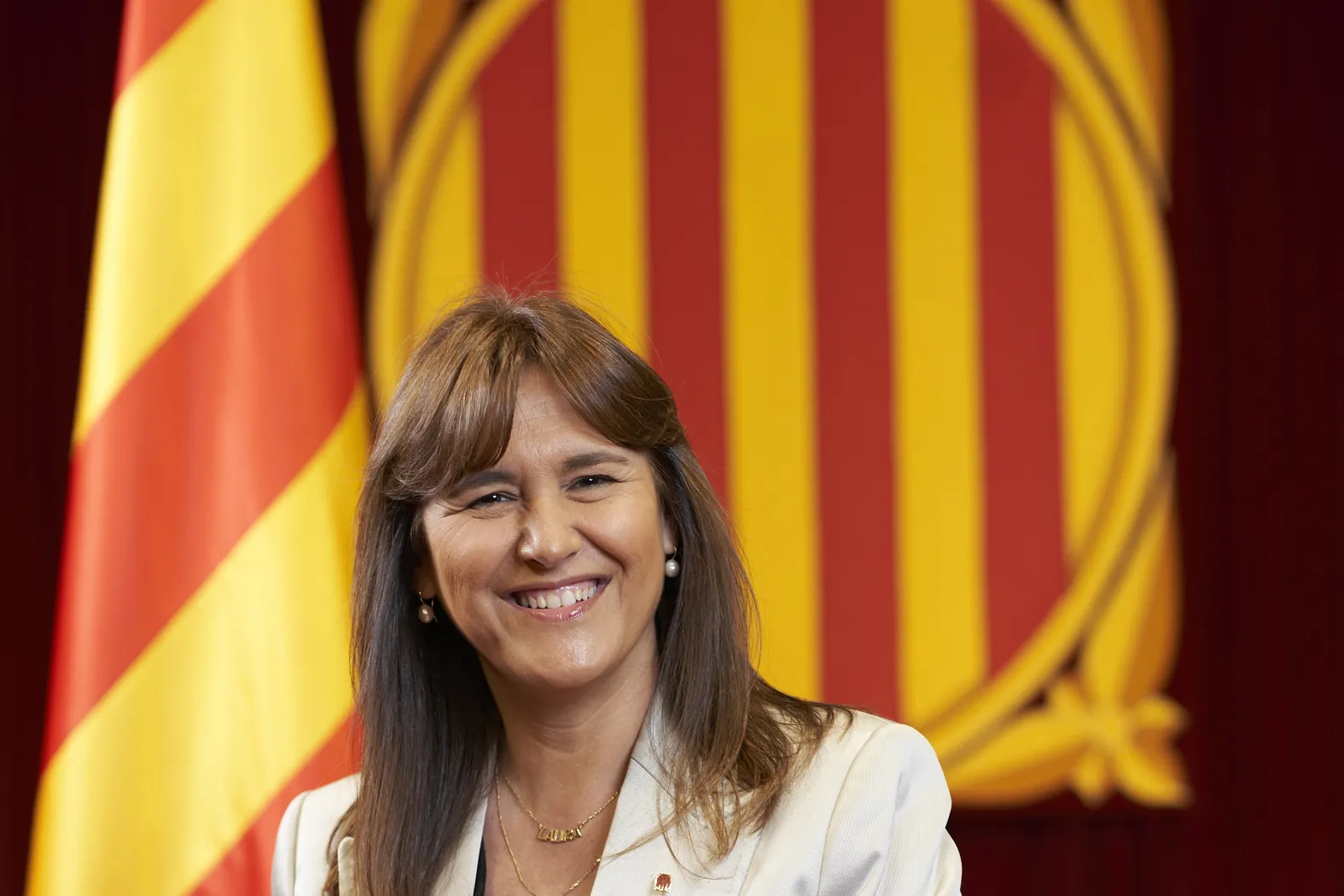 Laura Borràs sonríe en su retrato oficial como presidenta del Parlament, ante una bandera cuatribarrada y el emblema del Parlamento de Cataluña.
