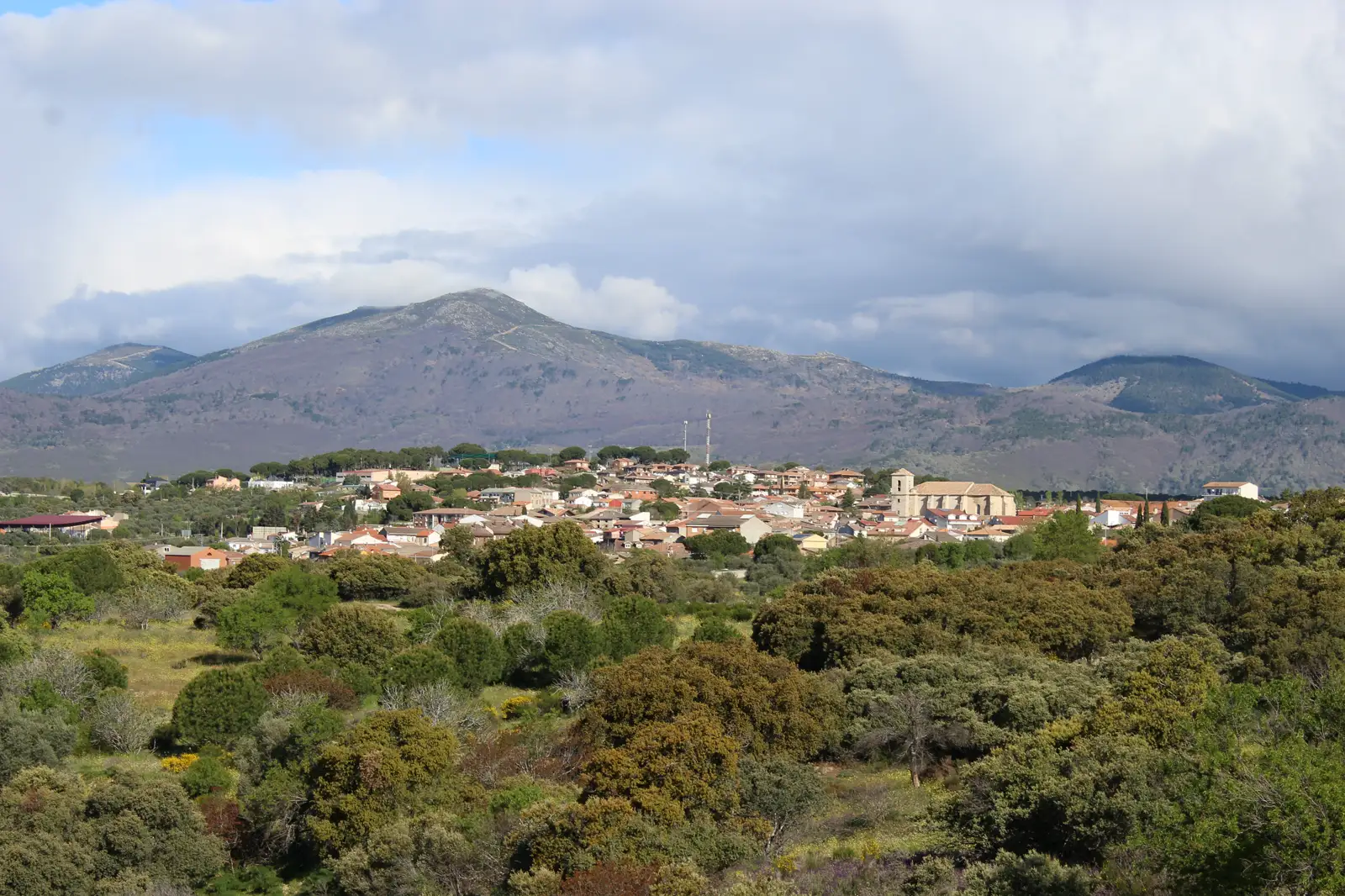 Vista de Cadalso de los Vidrios (Madrid) desde una ladera: en primer plano un monte de encinas y jaras, detrás el caserío de tejados rojizos con la torre de la iglesia, y al fondo las lomas boscosas de la sierra bajo un cielo de nubes.