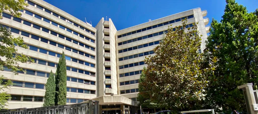 Fachada del edificio de la sede del Instituto Nacional de la Seguridad Social en Madrid, un bloque de oficinas de ocho plantas en hormigón claro con hileras de ventanas, rodeado de árboles y con cielo azul despejado.