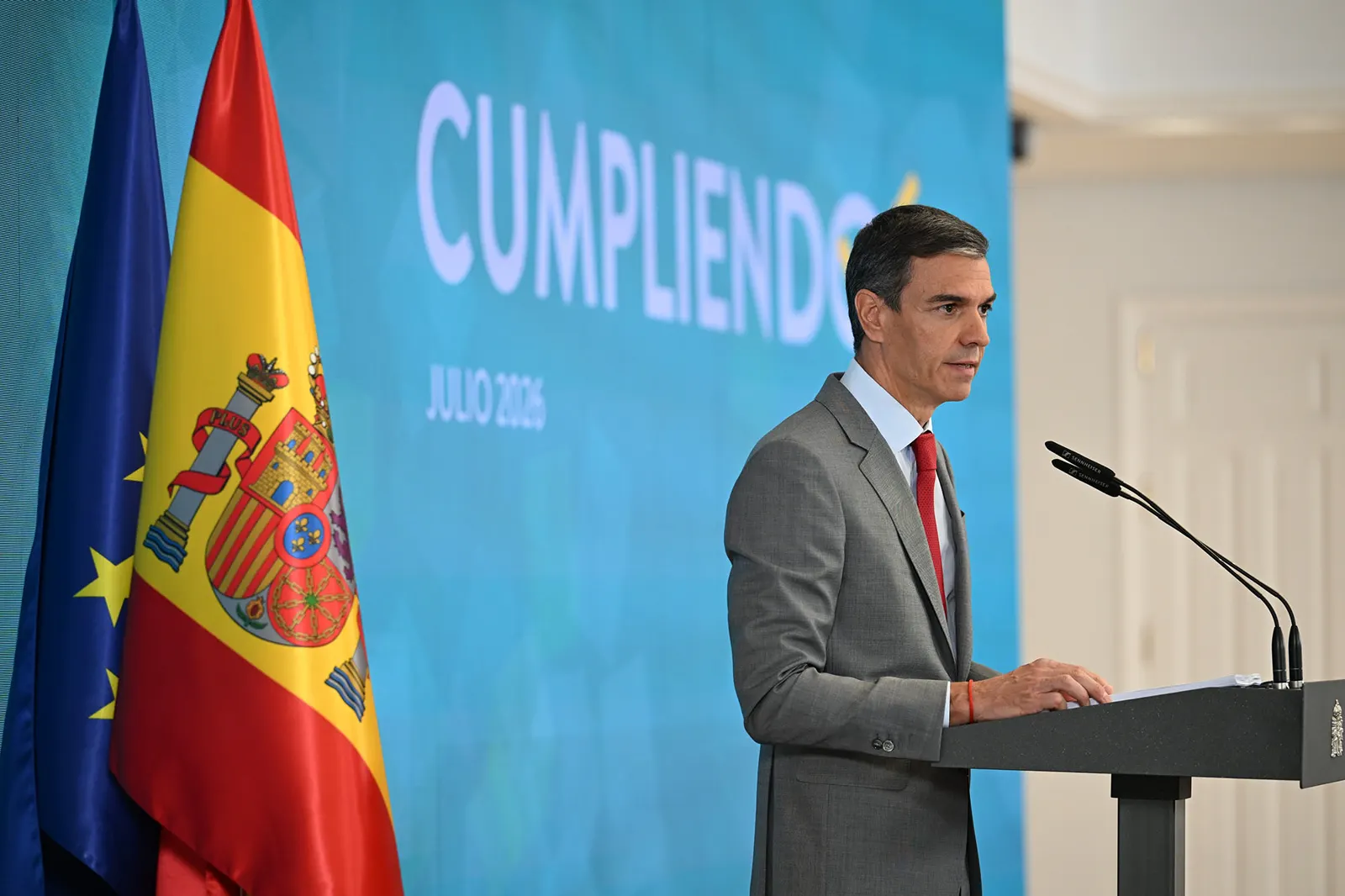 El presidente del Gobierno, Pedro Sánchez, de perfil ante un atril gris con el escudo de España, hablando a dos micrófonos con las banderas de España y de la Unión Europea a su izquierda y un panel azul de fondo con la palabra «Cumpliendo» y la fecha «julio 2026».