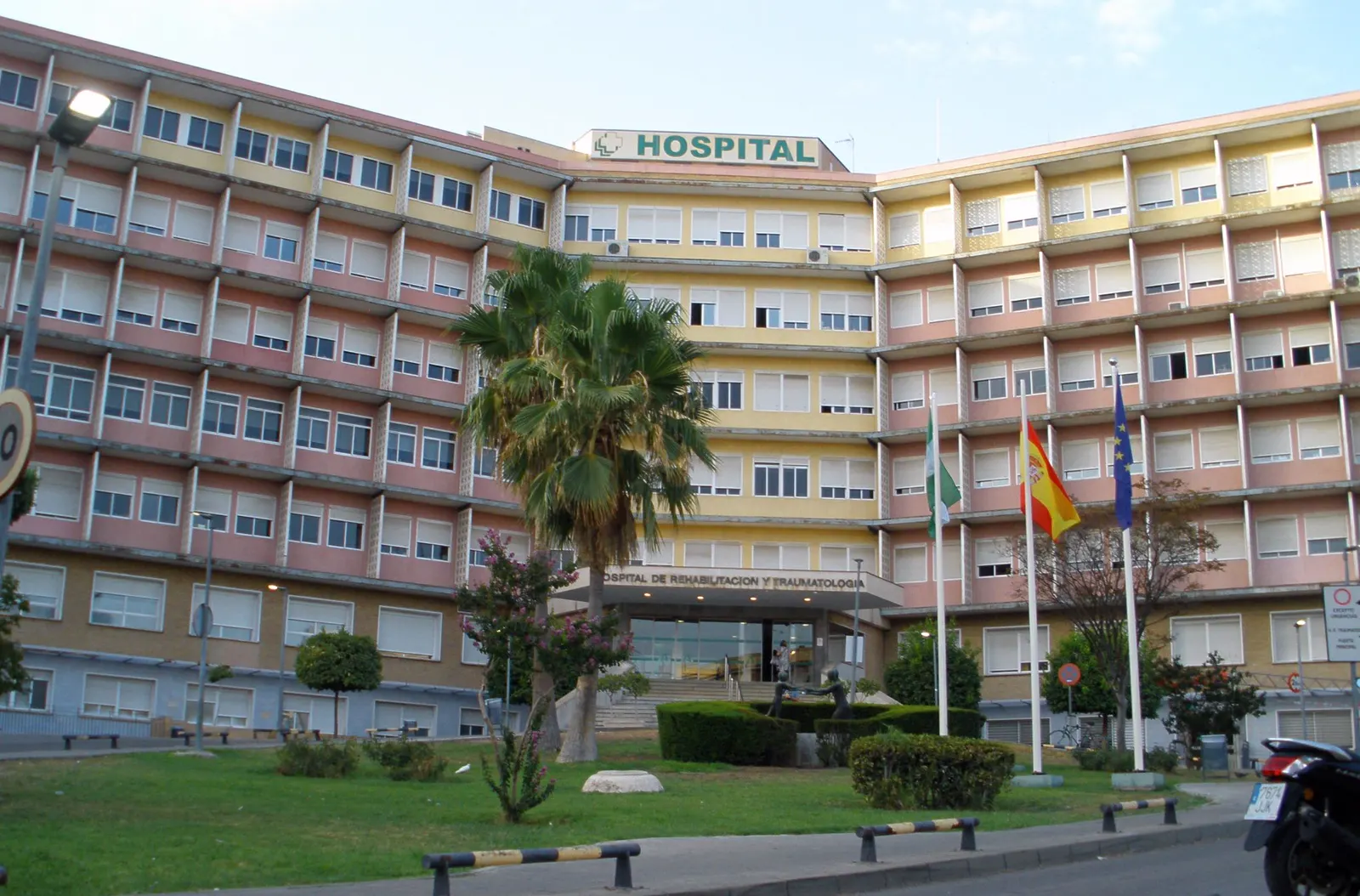 Fachada de un edificio hospitalario de seis plantas con el rótulo verde «HOSPITAL» sobre la entrada, una palmera en el jardín delantero y tres banderas —andaluza, española y europea— en sus mástiles.