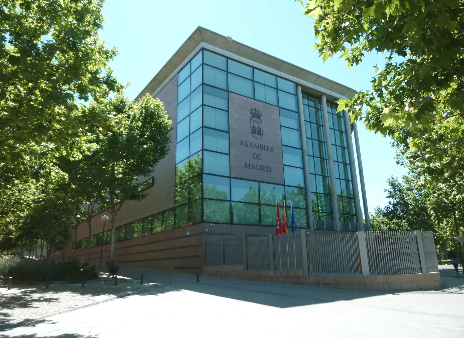 Fachada acristalada de la sede de la Asamblea de Madrid, con el escudo de la Comunidad y el rótulo «Asamblea de Madrid», entre árboles, en un día soleado.